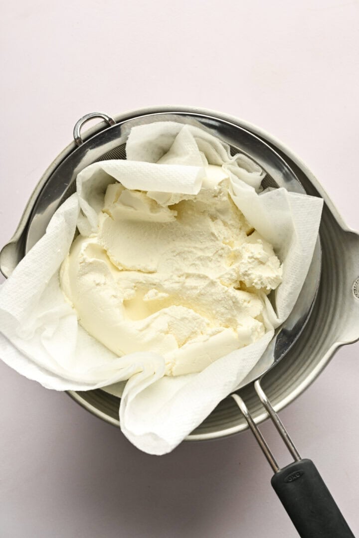 Ovanifrån av ricotta som rinner över ett durkslag insvept i pappershandduk.