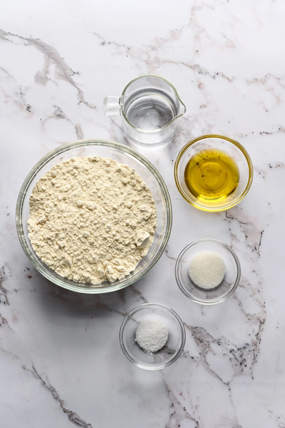 ovanifrån av mjöl, vatten, olivolja, salt och socker i skålar.
