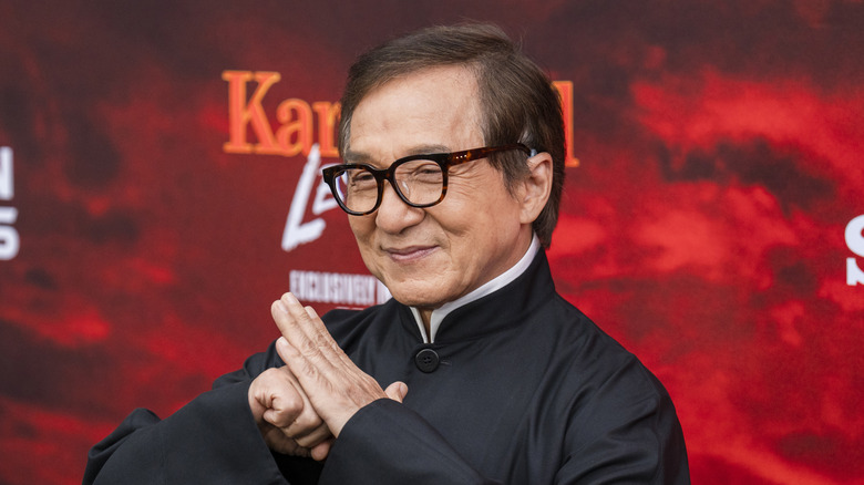 Jackie Chan framför en röd bakgrund med händerna ihop.