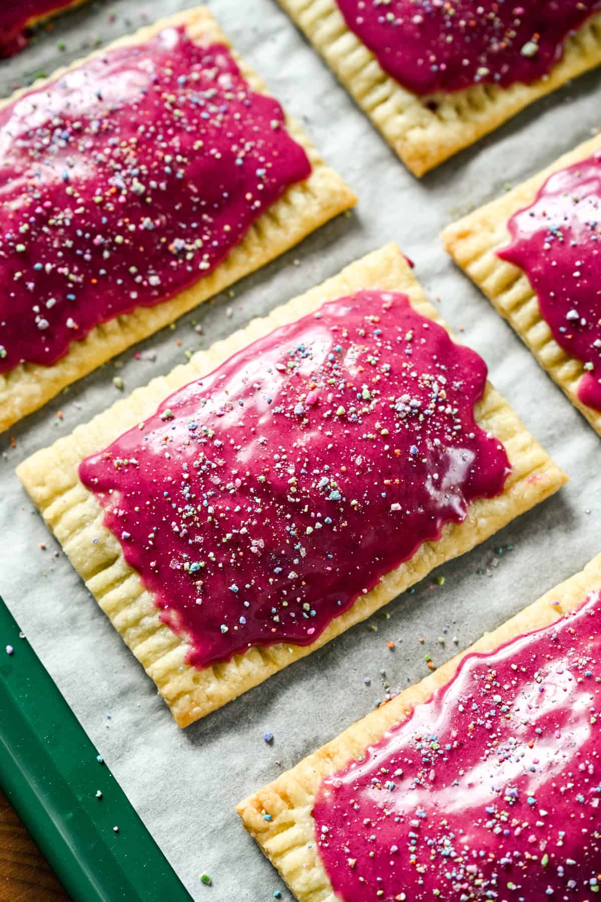 närbild av hemgjorda blåbärspoptårtor med ljust rosa glasyr och strössel på en plåt.