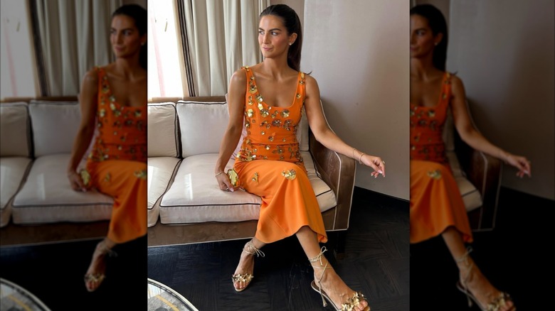 Kaitlan Collins sitter i kors på en vit och brun soffa. Hon är klädd i en orange klänning täckt av 3D-guldblommiga applikationer, klackar med remmar i guld och minimala guldsmycken. Hennes hår ligger bakåt bak axlarna