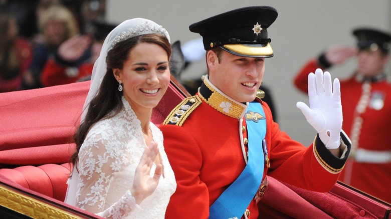Kate Middleton sitter i en vagn bredvid prins William. Hon är klädd i en spetsvit höghalsad brudklänning med diadem och slöja. William är klädd i röd kaptensuniform och fodermössa