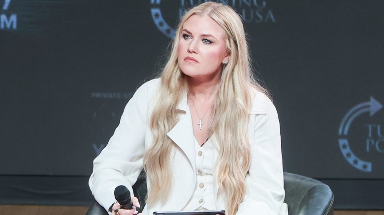 Erika Kirk sitter vid ett Turning Point USA-evenemang. Den här bilden visar hennes ultralånga blonda förlängningar. Hon är en kvinna med ljus hy, och är klädd i en vit kostymjacka och matchande väst.