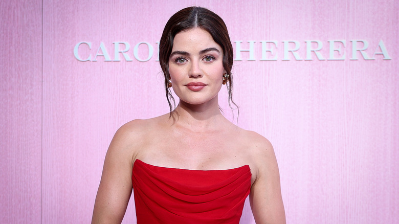 Lucy Hale bär en röd axelbandslös klänning medan hon poserar framför en rosa bakgrund