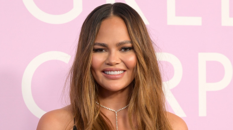 Chrissy Teigen ler vid ett evenemang på röda mattan iförd ett diamanthalsband mot en rosa bakgrund.