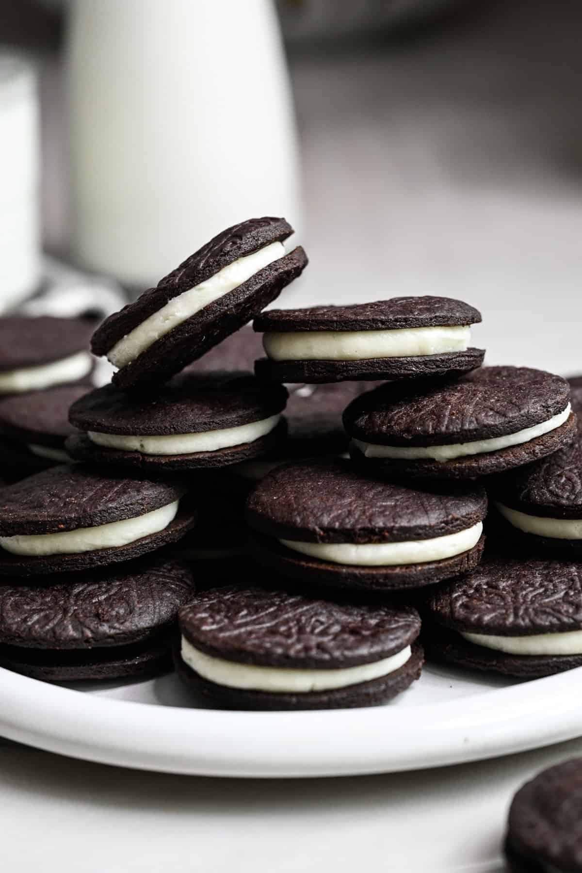 Närbild från sidan av hemgjorda oreos staplade på en vit platta med ett glas mjölk bakom.