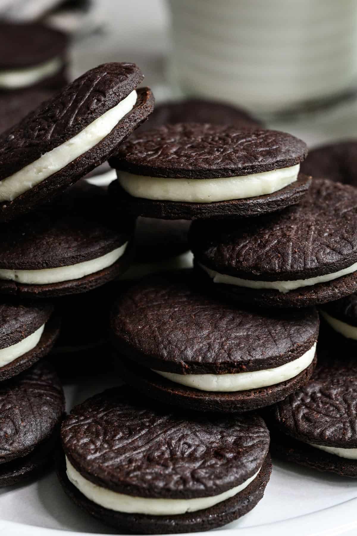 Närbild från sidan av hemgjorda oreos staplade på en vit platta med ett glas mjölk bakom.