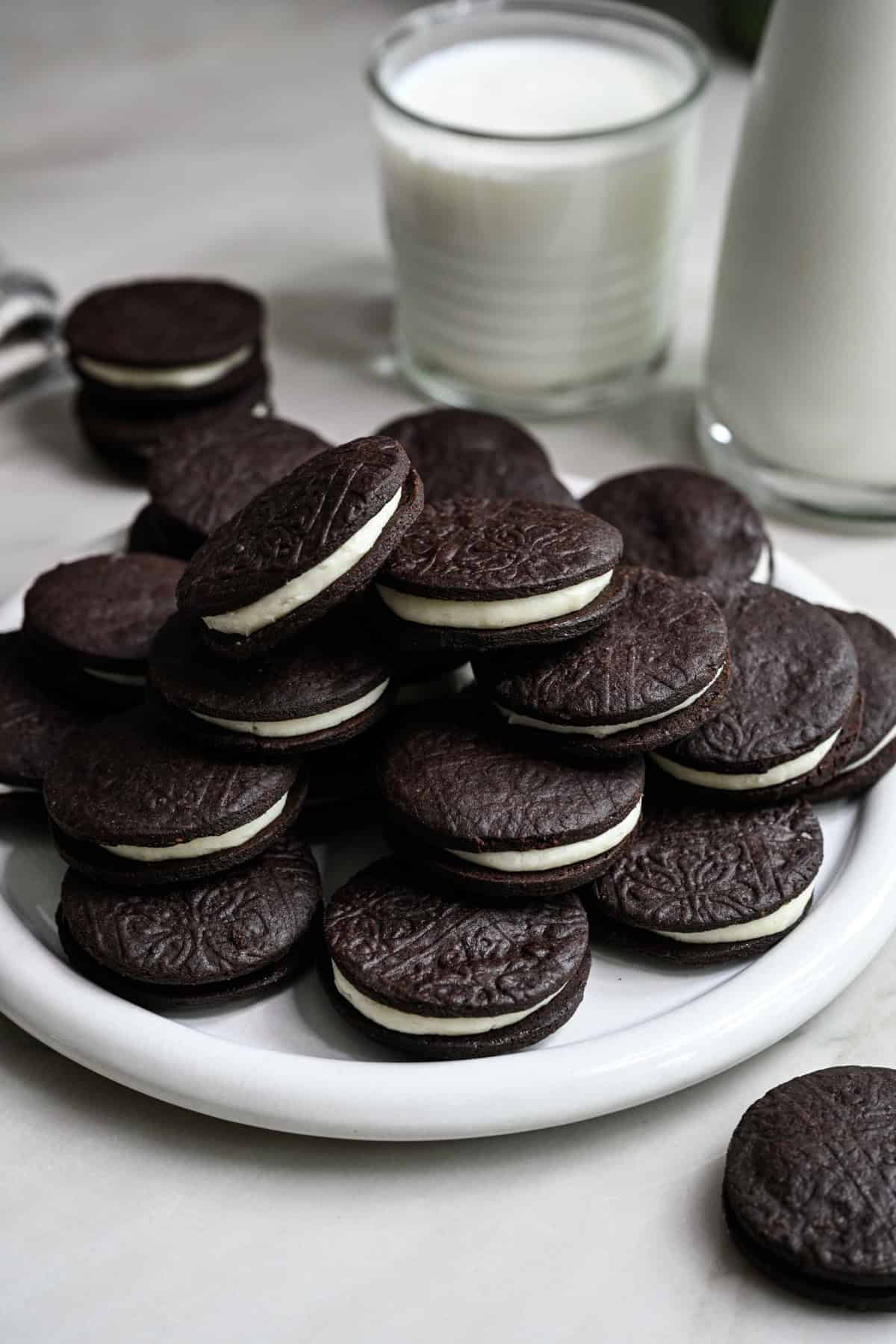 Närbild från sidan av hemgjorda oreos staplade på en vit platta med ett glas mjölk bakom.