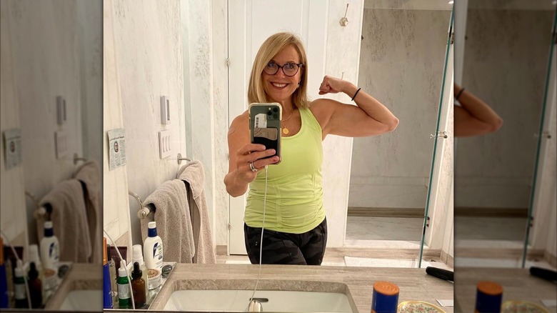 Katie Couric poserar för en spegelselfie när hon visar upp bicepsmuskeln. Hon har på sig en grön träningstank och kamouflageleggings.