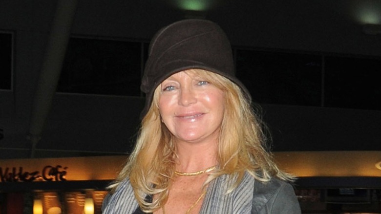 Goldie Hawn ler iklädd en svart hatt och grå jacka med guldhalsband.