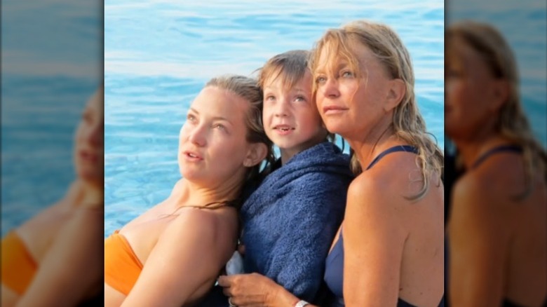 Kate Hudson, sonen Ryder och Goldie Hawn poserar och tittar uppåt vid vattnet i badkläder.