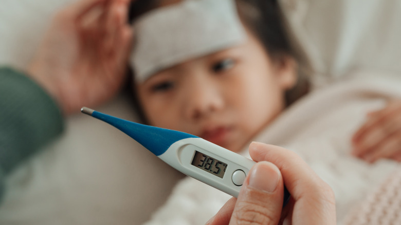 En hand som håller en digital termometer medan ett barn med feber lägger sig
