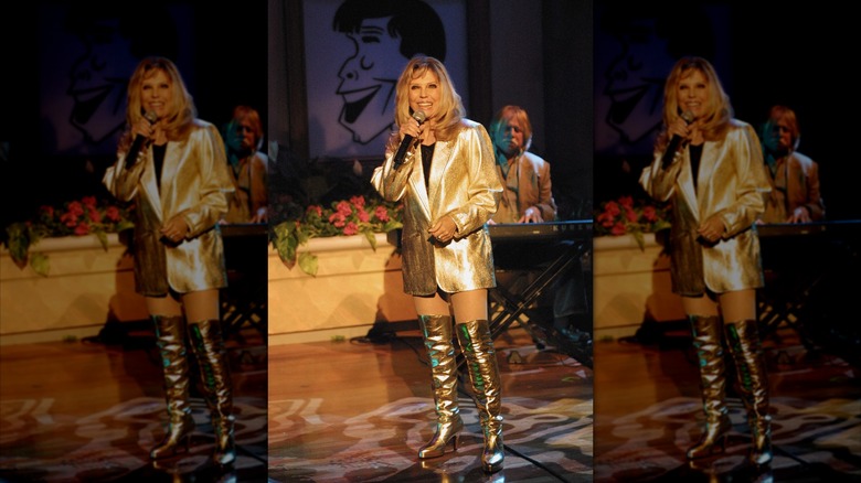 Nancy Sinatra uppträder på scen med en mikrofon iklädd knähöga stövlar i guld och en guldkavaj.