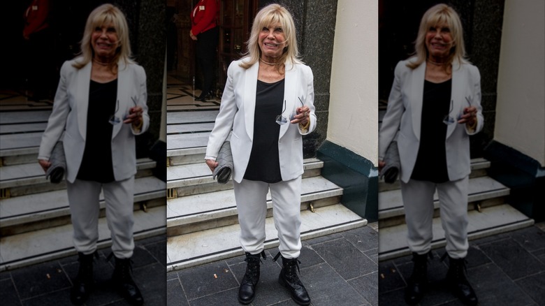Nancy Sinatra fotograferade utanför med sina glasögon och iklädd vita byxor en kavaj med svart skjorta och stridsstövlar.