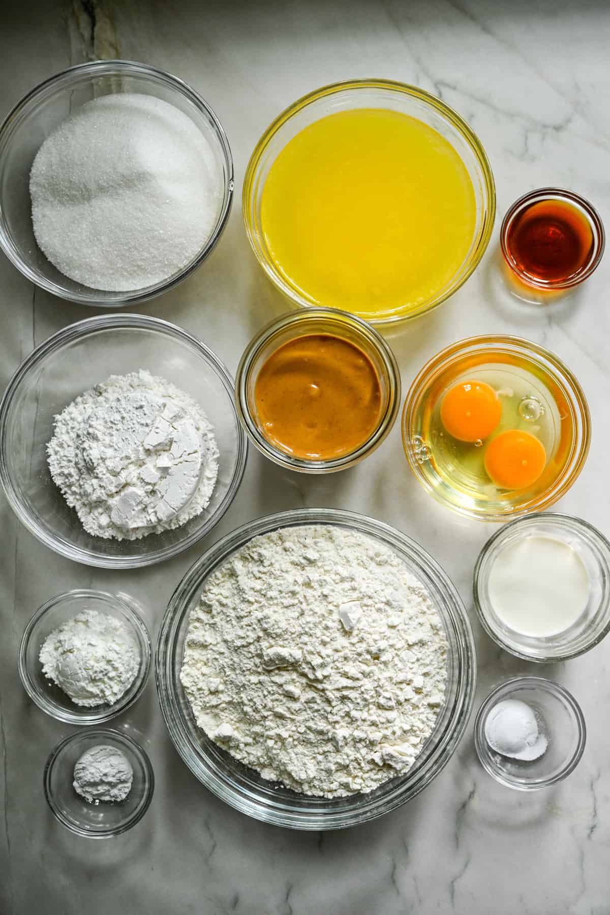 Ovanifrån av ingredienser för hemgjorda nötsmör i separata förberedande skålar på vit bänkskiva.