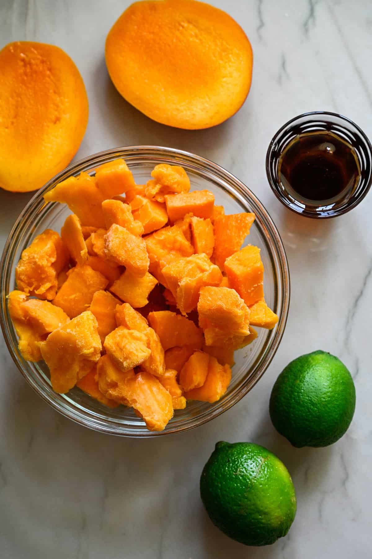 ovanifrån av tärnad mango i en skål, 2 limefrukter och agavesirap på bänkskivan.
