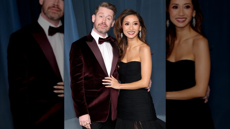 Macaulay Culkin i vinröd Paul Smith-kostym i mocka poserar bredvid Brenda Song i en SS23 Carolina Herrera-klänning.