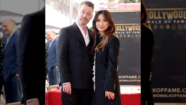 Macaulay Culkin i svart kostym står bredvid Brenda Song i en lång svart trenchcoat.