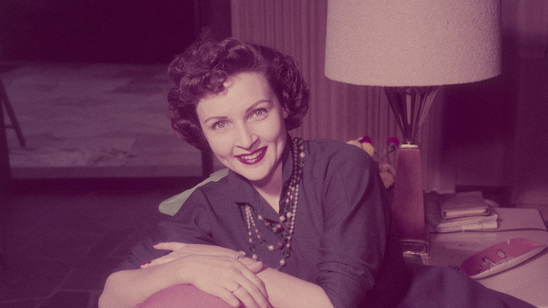 Betty White, amerikansk skådespelerska och komiker, ler medan hon sitter med armarna korsade över en kudde på ryggstödet på en soffa i ett vardagsrum, cirka 1955