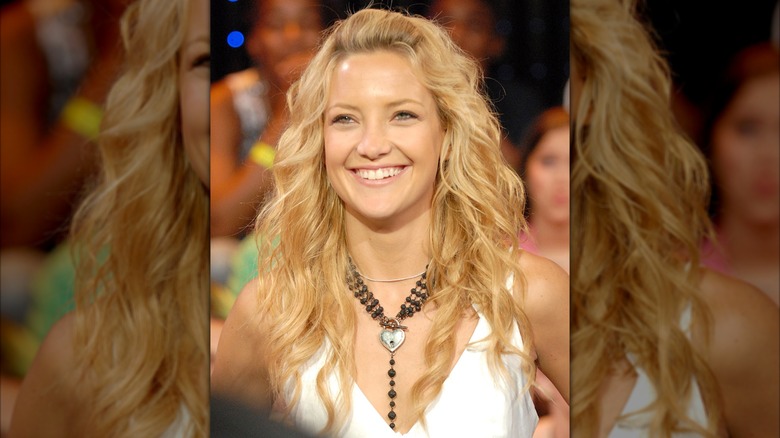 Kate Hudson i en vit beskuren väst med ett svart chunky halsband och håret i lösa vågor med en pangpuff.