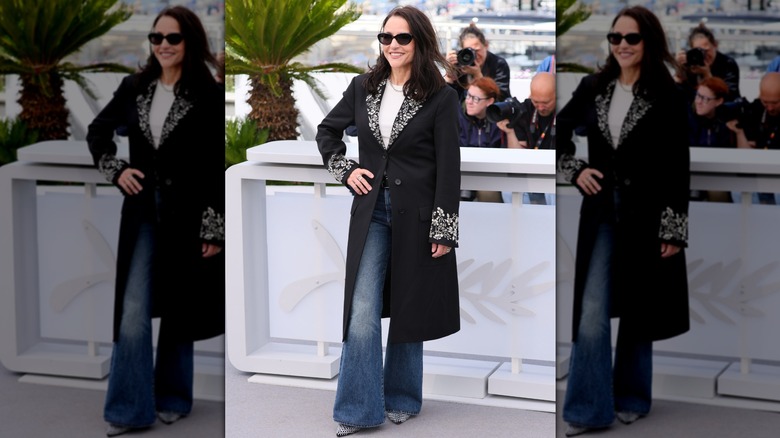 Julia Louis-Dreyfus poserar på ett evenemang med handen på höften, iklädd utsvängda jeans, en svart kappa och mörka solglasögon.
