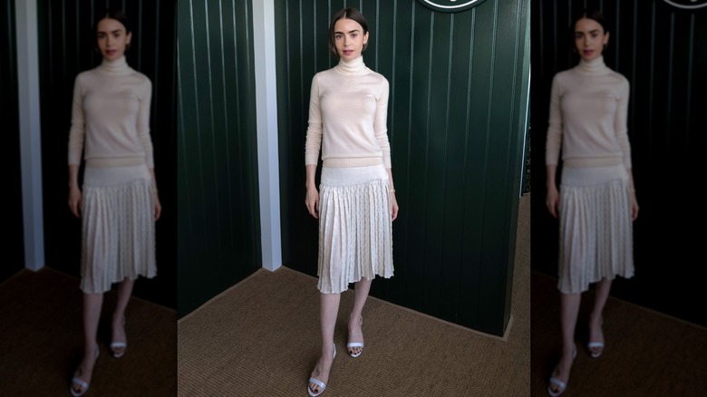 Lily Collins iklädd en krämfärgad turtleneck-topp och en vit veckad kjol med vita sandaler på ett event.