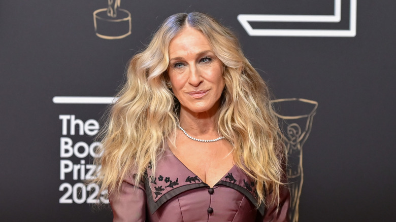 Sarah Jessica Parker poserar på ett event iklädd en rödbrun klänning med vågigt hår och ett delikat halsband.