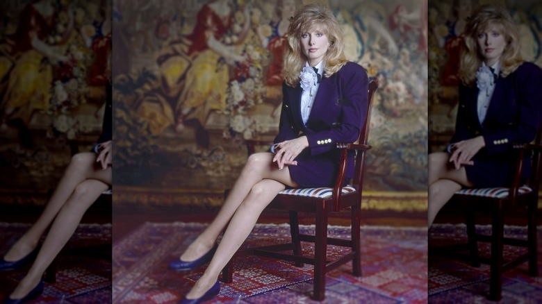 Morgan Fairchild sitter i en stol med korsade ben iförd en mörklila kjolkostym.