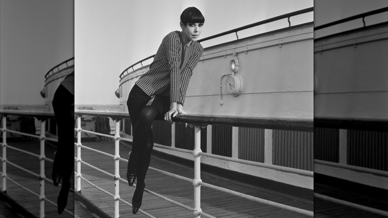 Peggy Moffitt poserar på räcket på ett skepp iförd en rutig jacka med svart kjol och strumpbyxor.