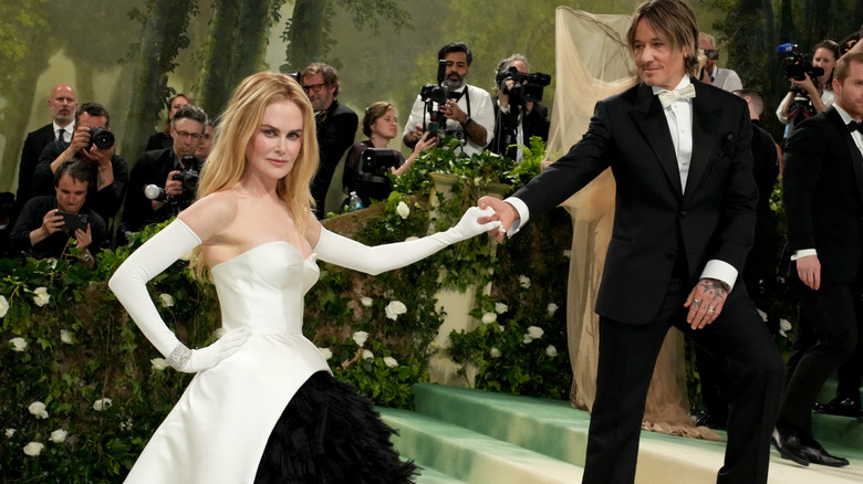 Nicole Kidman poserar i en vit axelbandslös klänning med vita operahandskar som tar Keith Urbans hand vid ett event 2024.