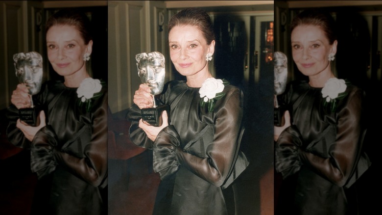 Audrey Hepburn poserar med en utmärkelse klädd i en svart klänning med skira ärmar och en vit rosaccent.
