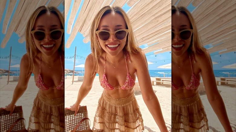 Camelia Somers bär solglasögon, en rosa blommig bikiniöverdel och skirt brun kjol på stranden.