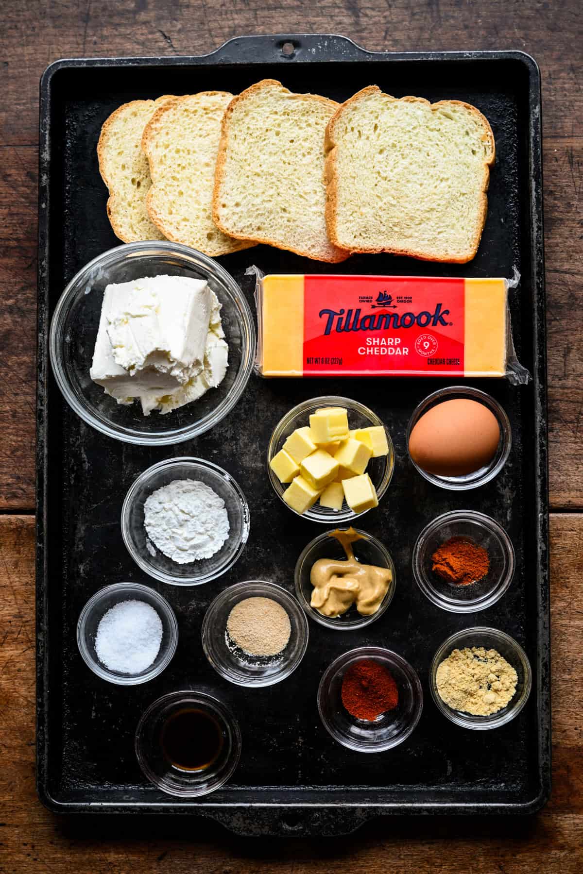 Vy ovanifrån av ostdrömmar ingredienser på en mörk plåt: bröd, färskost, ett block av Tillamook skarp cheddar, tärning av smör, ett ägg, senap och små skålar med kryddor.