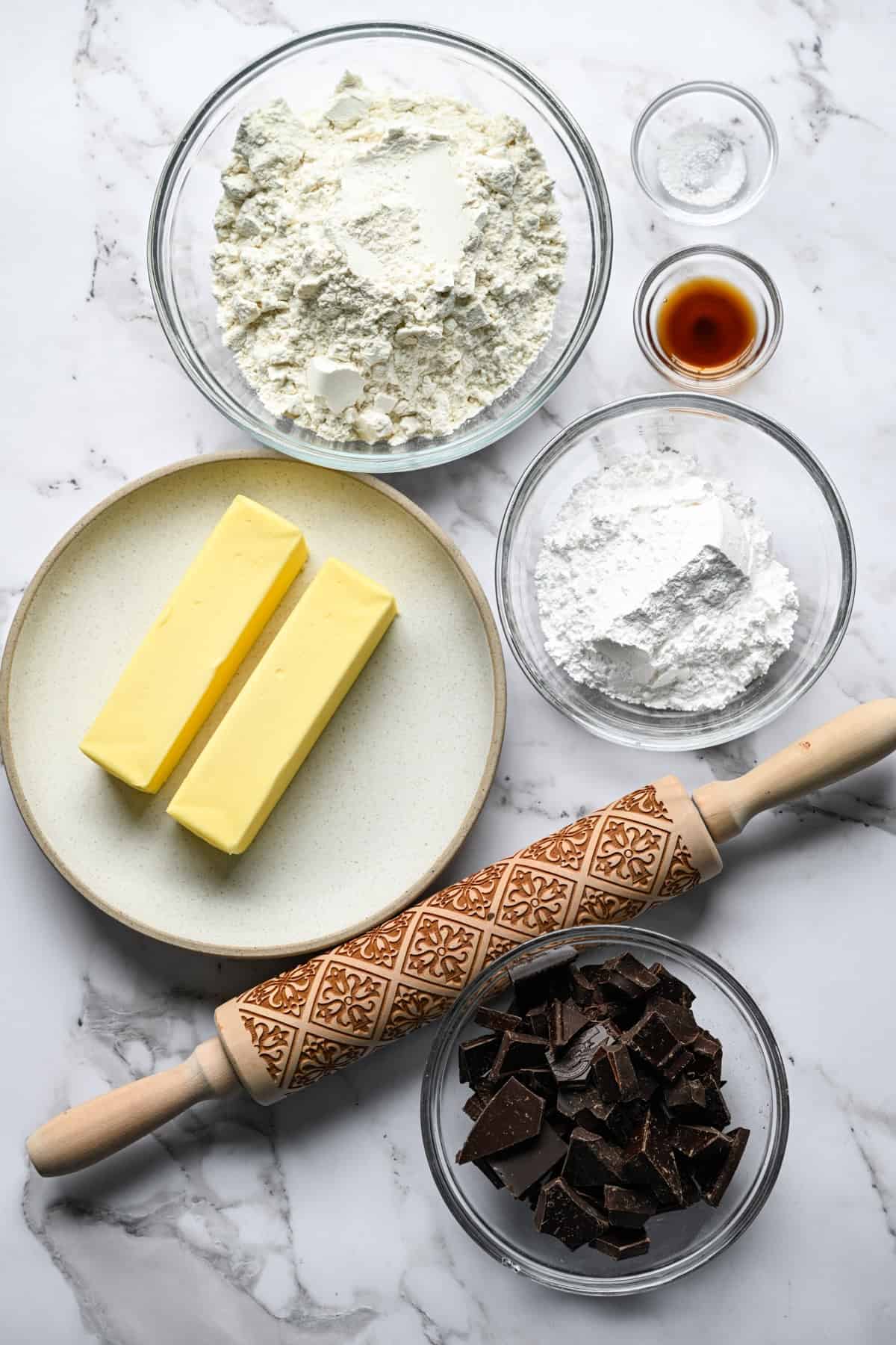 Glasskålar med mjöl, strösocker, hackad mörk choklad, vanilj och salt på en marmorbänk med två smörpinnar på en tallrik och en präglad träkavel.
