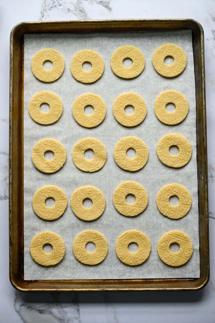 Tjugo obakade ringformade mördegskakor med ett präglat mönster uppradade i rader på en bakplåtspappersklädd plåt.