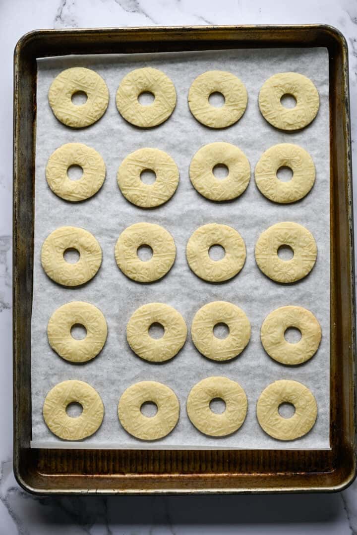 Tjugo bakade ringformade mördegskakor, blekt gyllene med ett präglat mönster, på en bakplåtspappersklädd plåt.