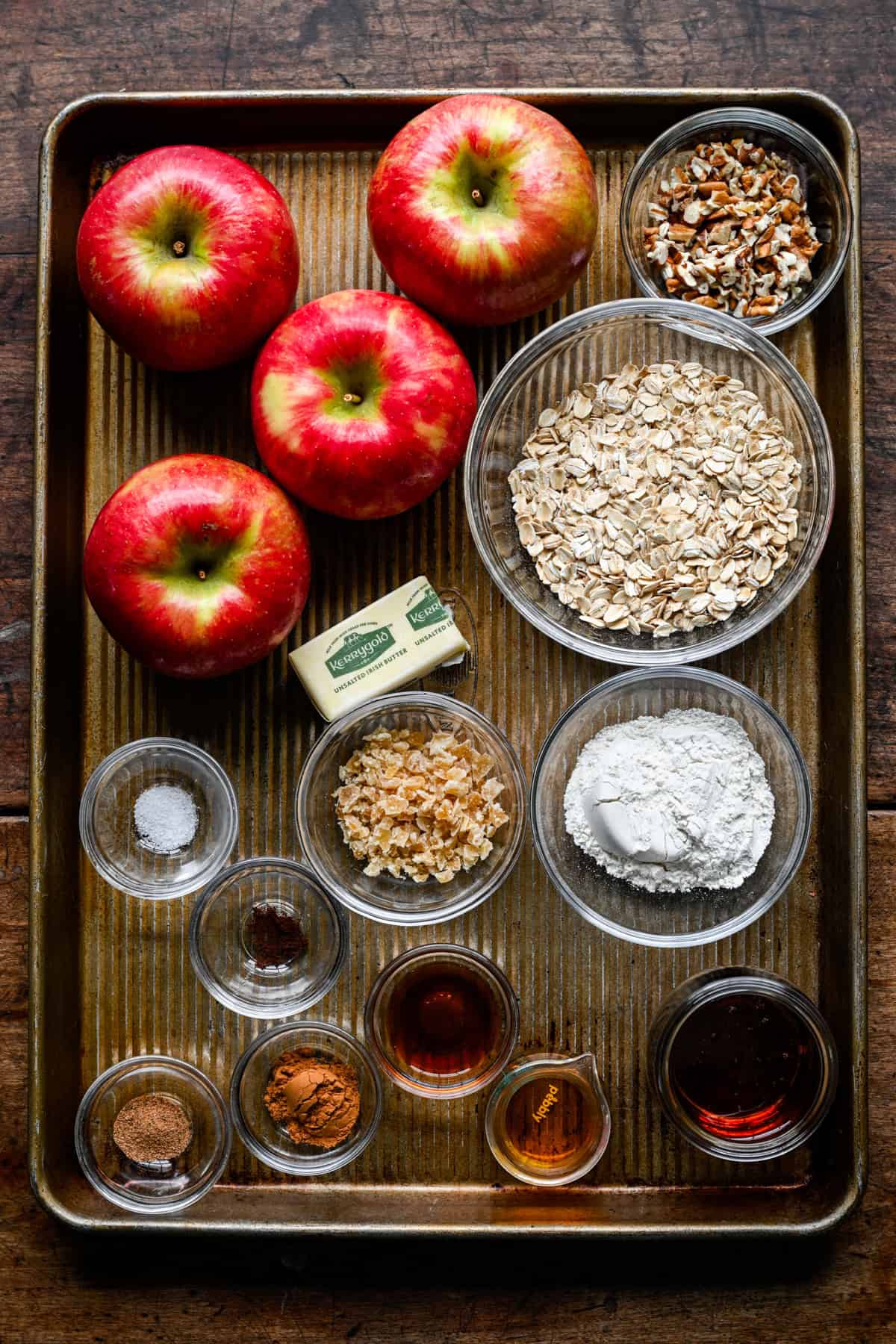 Ingredienser för bakade äpplen på en plåt: fyra honungsspridda äpplen, skålar med havregryn, hackade pekannötter, mjöl, kristalliserad ingefära, en smörstång, malda kryddor, lönnsirap och vanilj.