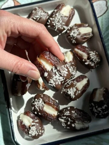 Närbild av en hand som lyfter en chokladtäckt dadel fylld med kokosnötsfyllning från en bricka.