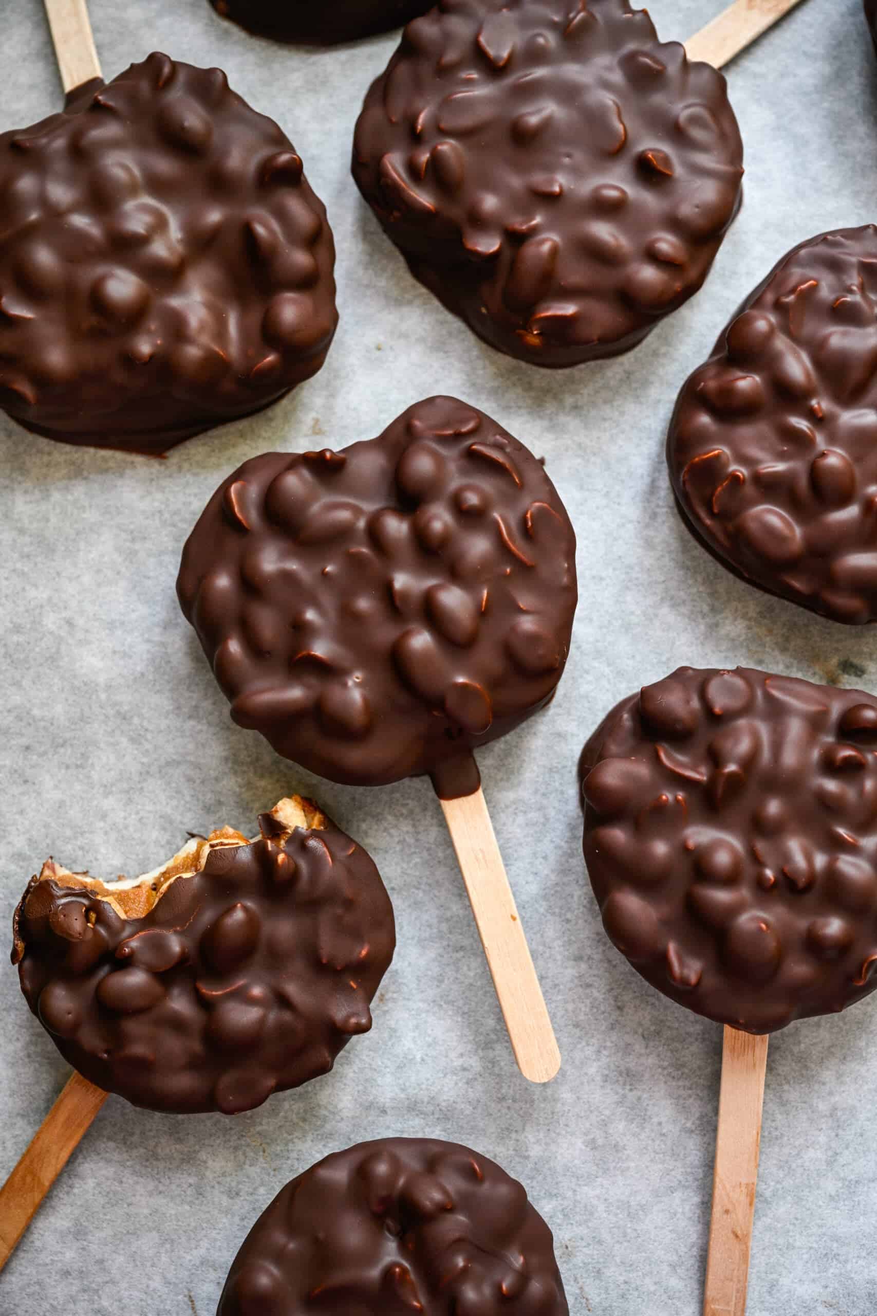 Vy ovanifrån av chokladtäckta äppelskivor på popsikelpinnar på bakplåtspapper, en med en tugga tagen ut.