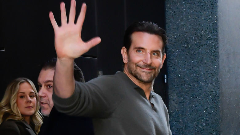 Bradley Cooper vinkar och ler när han går på en fullsatt gata