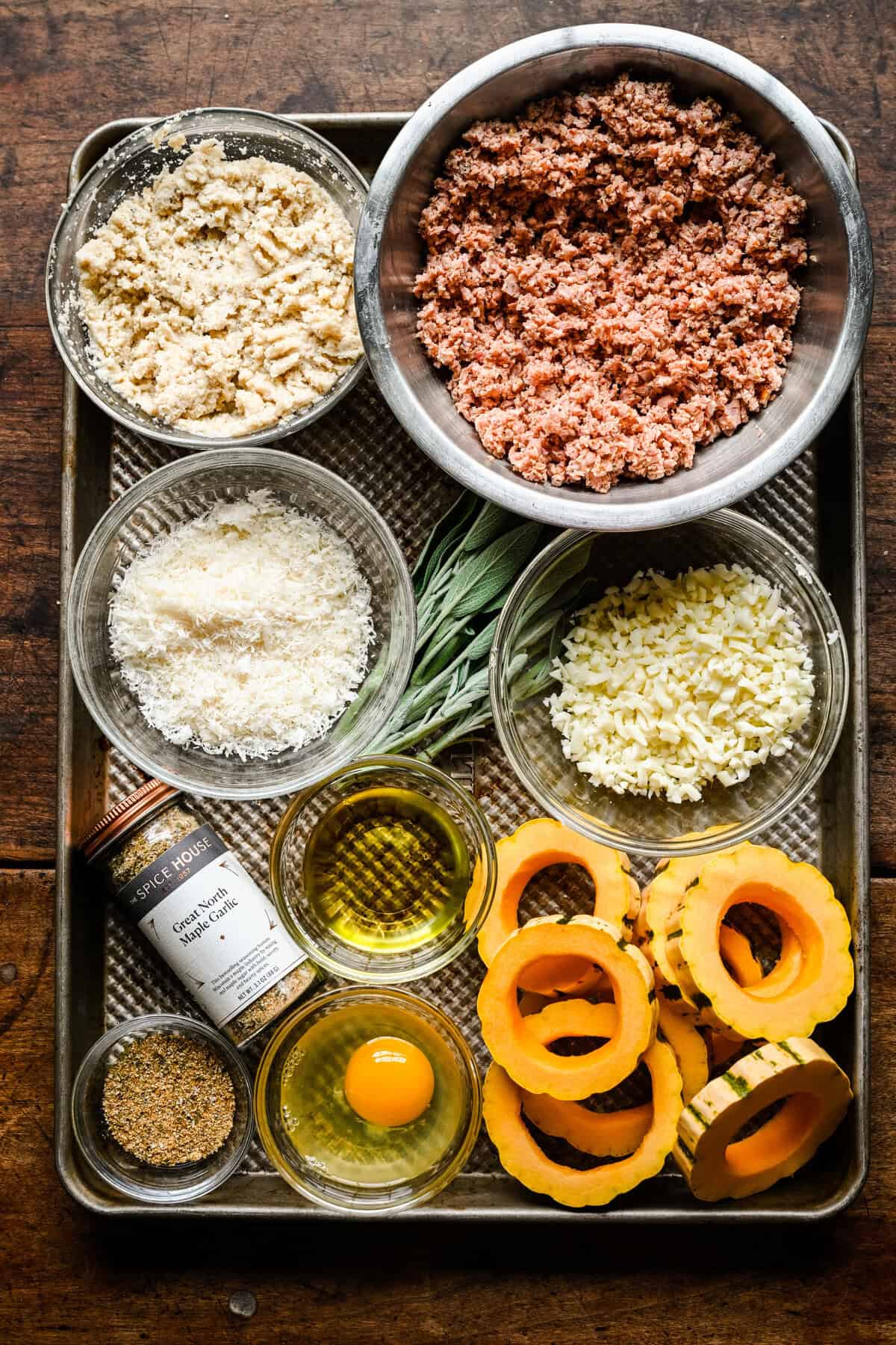 Vy ovanifrån av ingredienser på en plåt: mald korv, ströbröd, parmesan, mozzarella, färsk salvia, olivolja, ett ägg, Great North Maple Garlic-krydda och delikata squashringar.