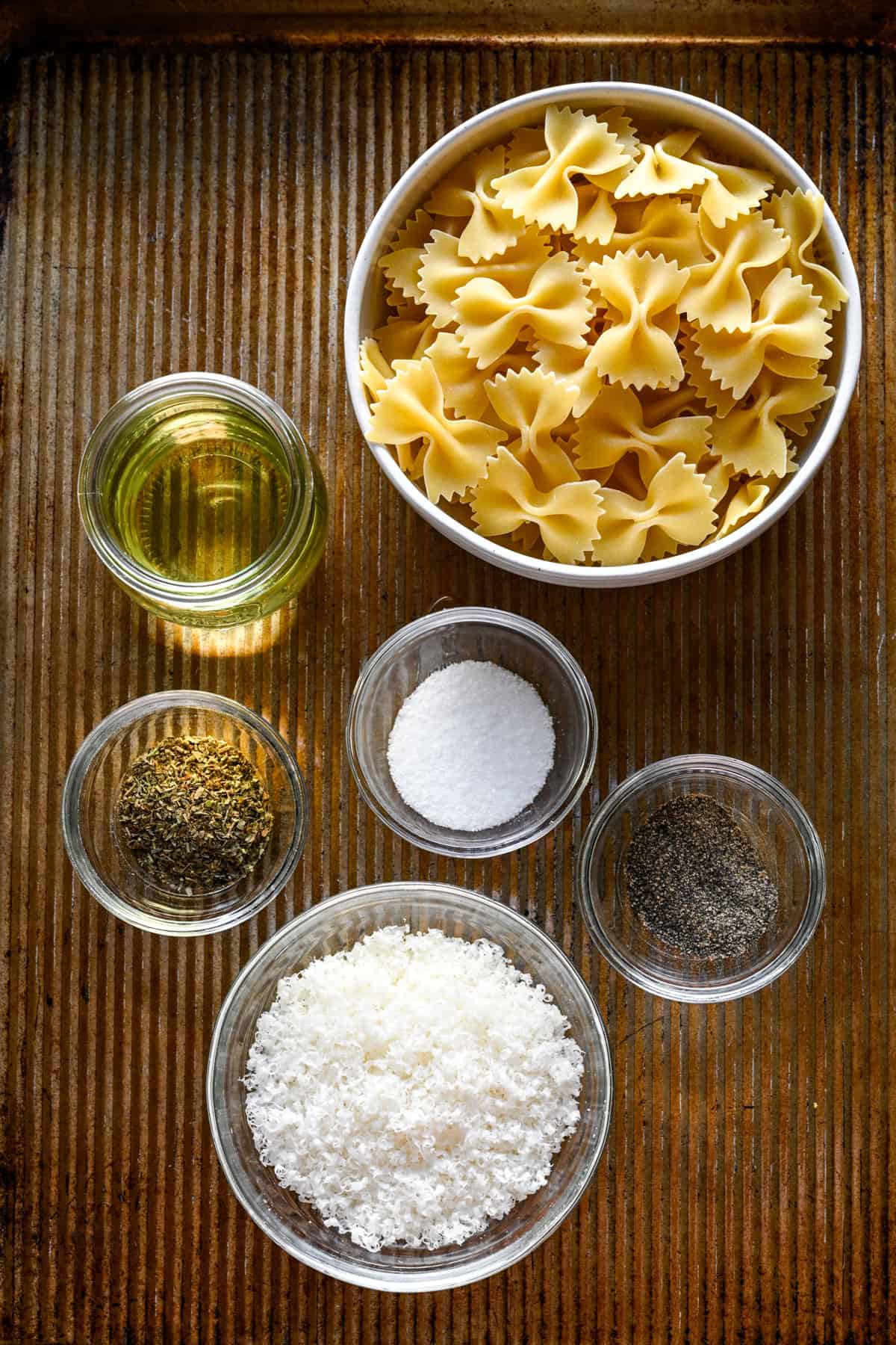 Ovanifrån av pastapuffingredienser på en plåt: fluga pasta, olja, torkade örter, salt, peppar och riven parmesan.