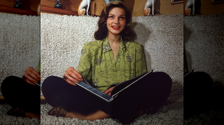 Lauren Bacall sitter i kors med en öppen bok i knät iförd en grön prydd blus och rött läppstift cirka 1945