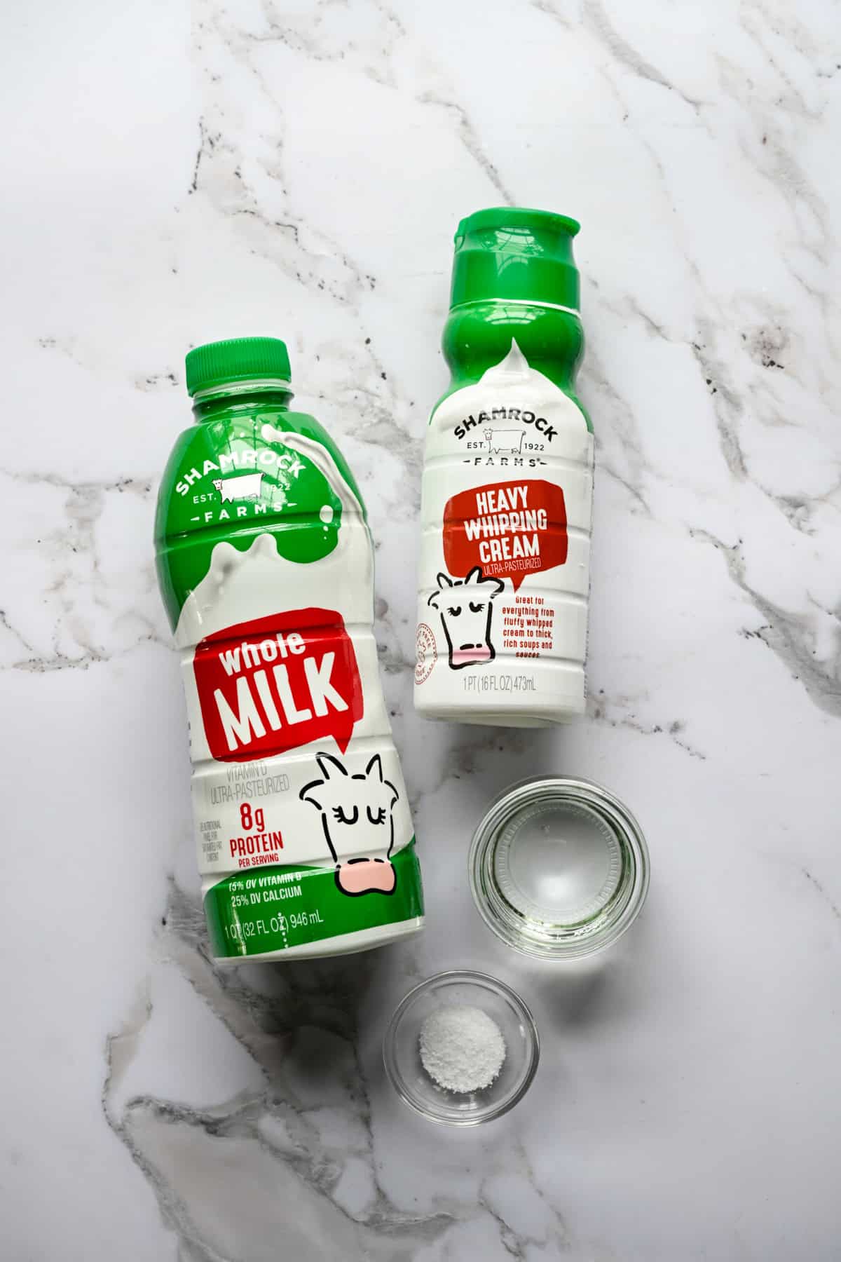 Vy ovanifrån av ingredienser till hemgjord keso: en flaska Shamrock Farms helmjölk, Shamrock Farms tung vispgrädde och små glasskålar med vit vinäger och salt på marmor