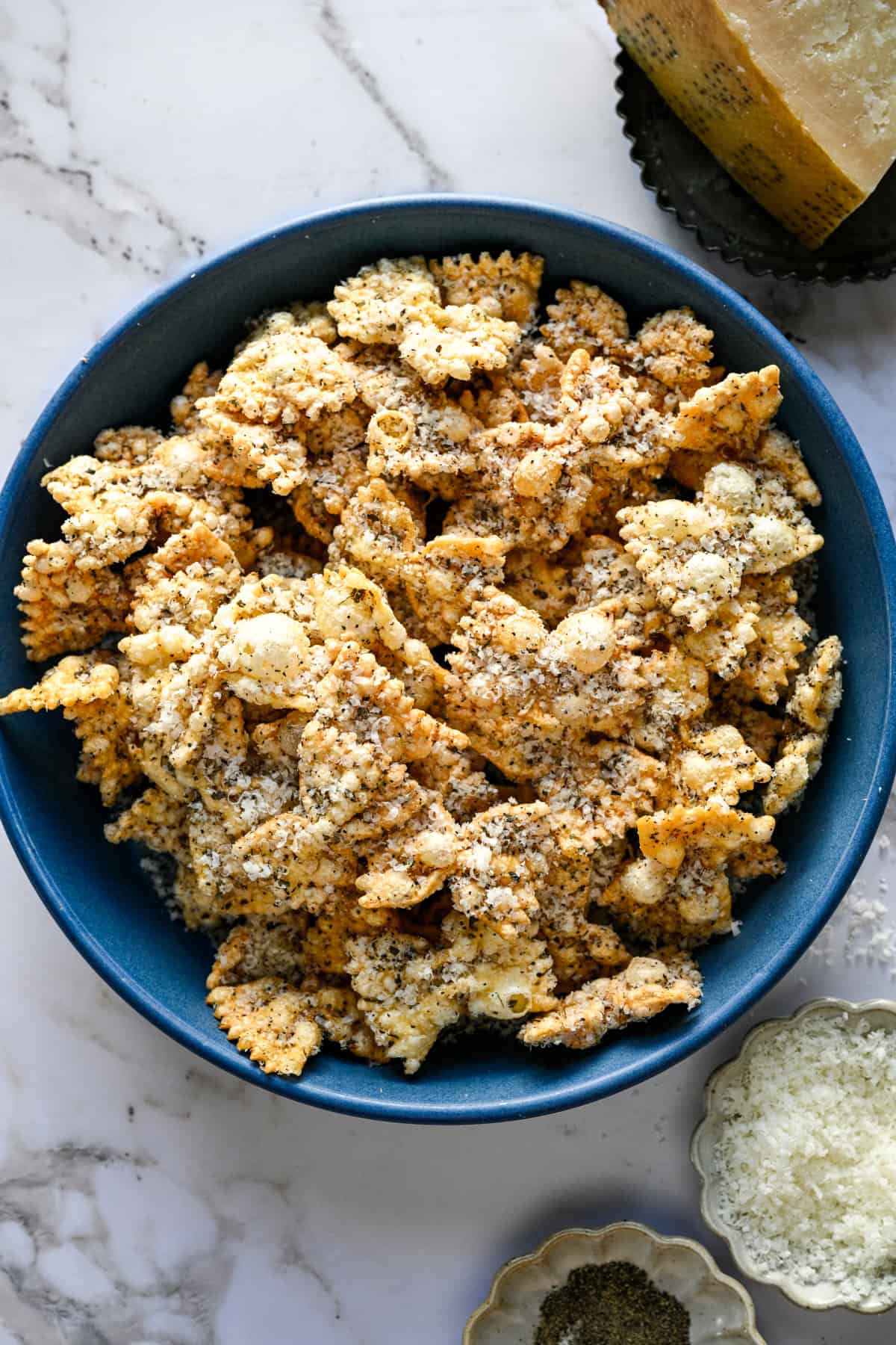 Ovanifrån av puffad pasta i en blå skål bredvid en klyfta parmesan och ett fat med peppar.
